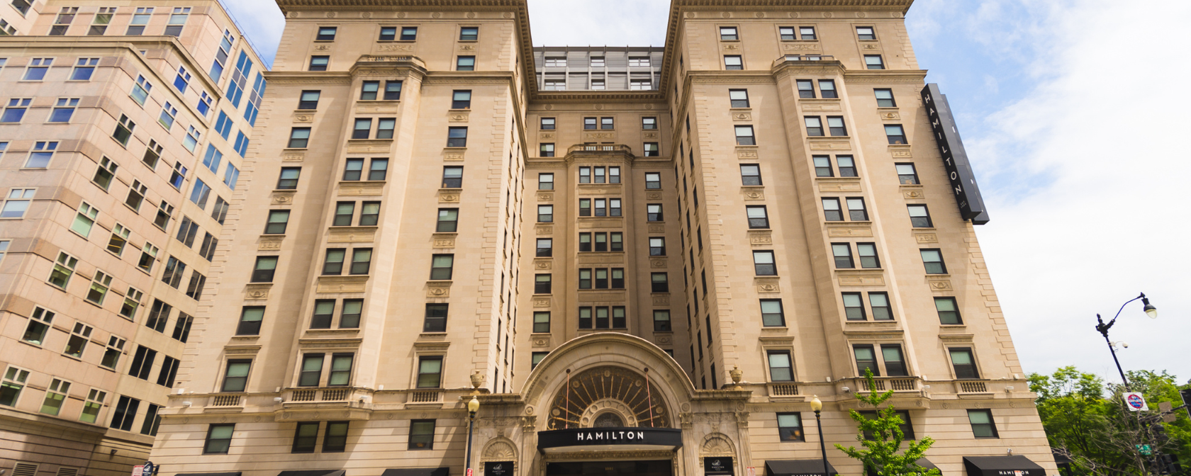 A grand, historic beige stone building named "Hamilton" with an ornate arched entrance, surrounded by modern structures under a partly cloudy sky.